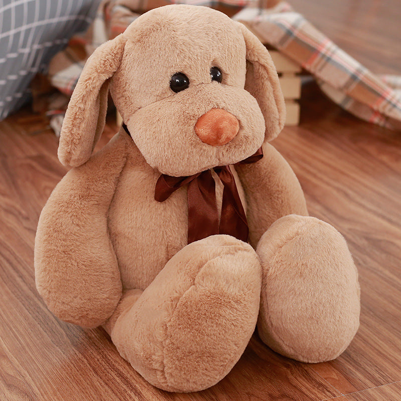Cute Dull Dog Doll is a light tan plush dog with floppy ears, dark eyes, a peach nose and brown bow